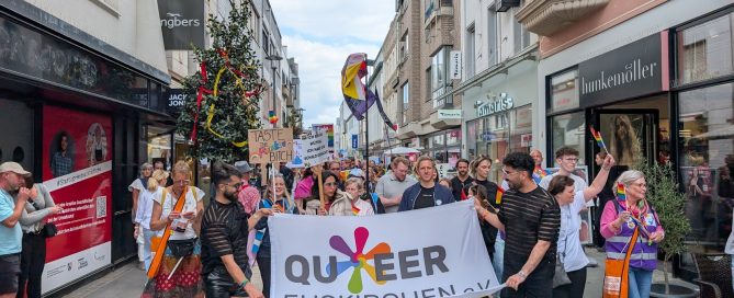 Menschen bei einer Parade mit einem Banner, das 'QUEER' zeigt, in einer Stadtstraße.