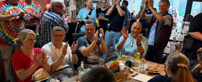 Eine Gruppe von Menschen applaudiert in einem Restaurant während einer Feier mit Regenbogenflaggen.